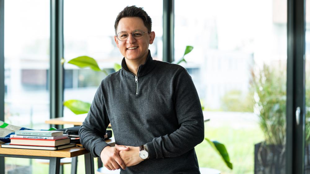 Professionelle Person steht lächelnd vor einem modernen Bürofenster und optimiert ihr LinkedIn-Profil.