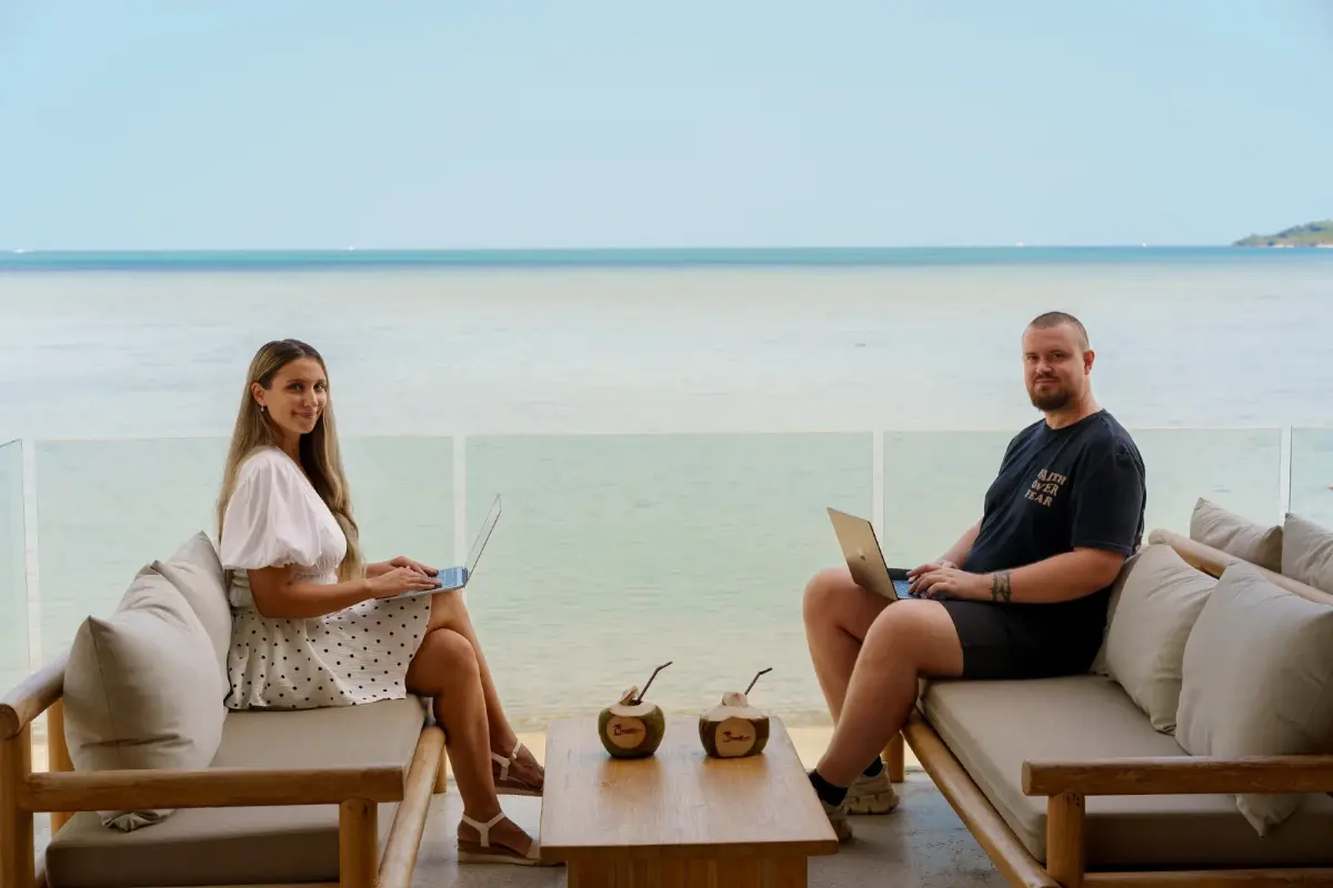 Paar arbeitet ortsunabhängig am Strand mit Laptops und Blick aufs Meer.