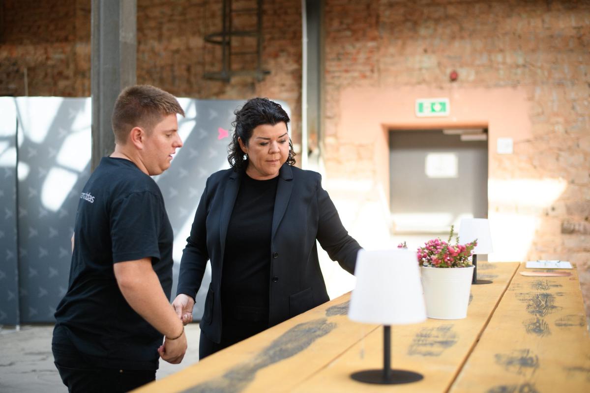 Firmenevent: Zwei Mitarbeitende sprechen an einem Stehtisch mit Lampe und Pflanze in einer Backsteinhalle.