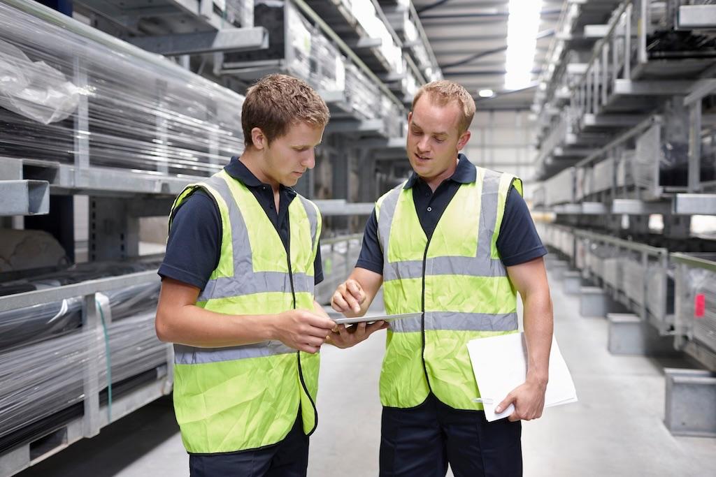 SAP-Logistik im Lager: Zwei Mitarbeiter in Warnwesten prüfen gemeinsam ein Tablet zwischen den Regalen.