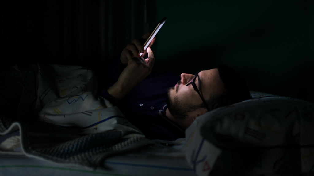 Mann liegt im Bett und schaut in sein Handy, während er über zwanghafte Masturbation nachdenkt.