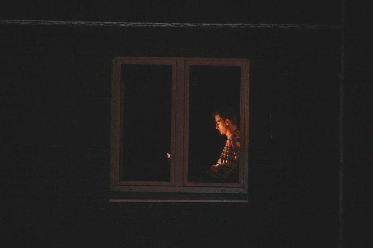 Mann sitzt nachts allein am beleuchteten Bildschirm hinter einem dunklen Fenster