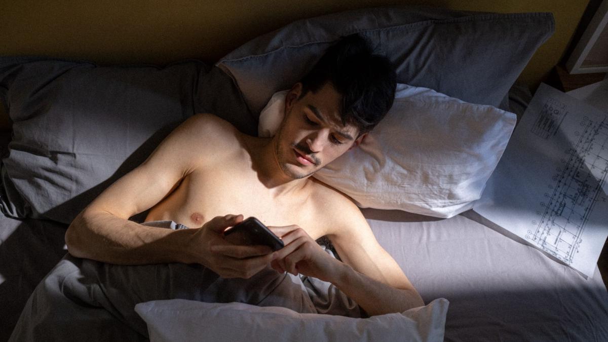 Ein Mann im Bett schaut auf sein Smartphone, das Thema ist Pornosucht und Krankheit.