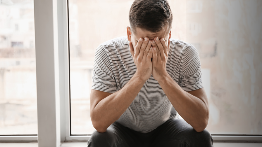 Mann sitzt im Innenraum und verdeckt sein Gesicht, kämpft mit seinen Emotionen beim Findom loswerden.