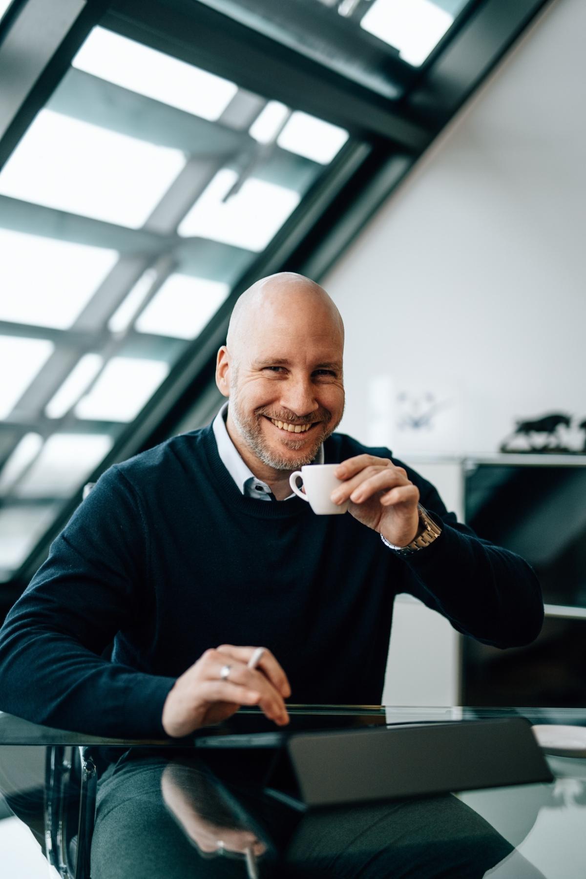 Mann mit Kaffee lächelt am Schreibtisch in einem modernen Büro.