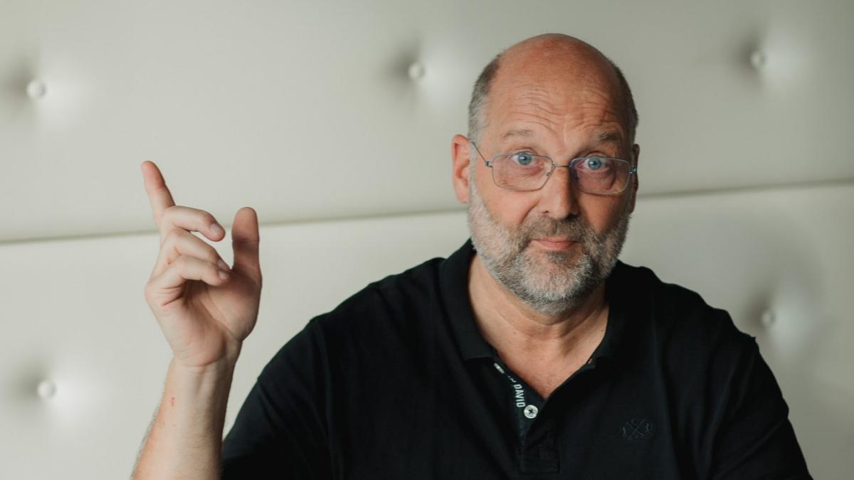 Ein älterer Mann im schwarzen Hemd zeigt nach oben; bKV-Leistungen unterstützen Mitarbeiter im Alltag.