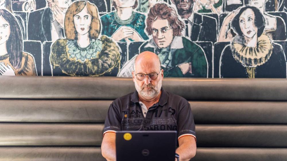 Ein Mitarbeiter sitzt mit einem Laptop vor einer Wand mit gezeichneten Porträts; bKV-Leistungen unterstützen Beschäftigte im Alltag.