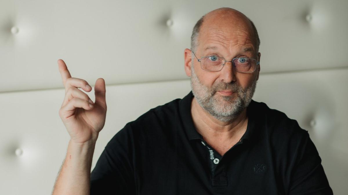 Ein älterer Mann mit Brille erklärt etwas mit erhobenem Zeigefinger vor einer hellen Polsterwand zur betrieblichen Krankenversicherung.