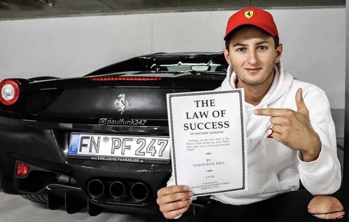 Junger Mann zeigt auf das Erfolgsgesetz, sitzt vor einem Ferrari.