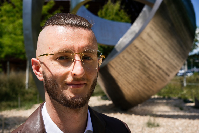 Junger Mann im Sonnenlicht mit Brille vor einer metallischen Skulptur, die das Thema Manifestieren symbolisiert.