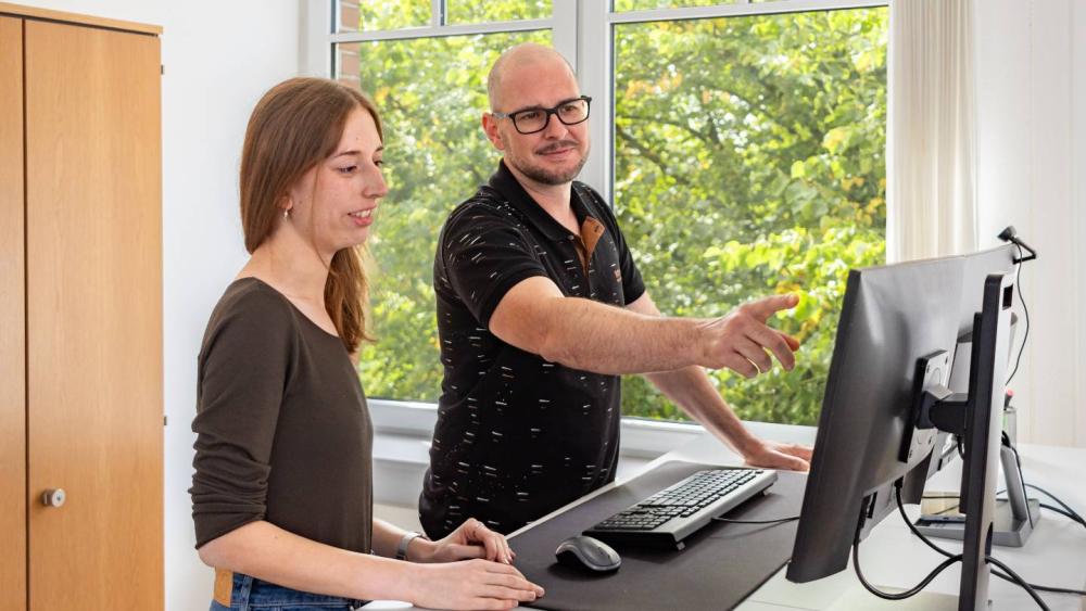 Zwei Personen diskutieren über KI-Sichtbarkeit an einem PC.
