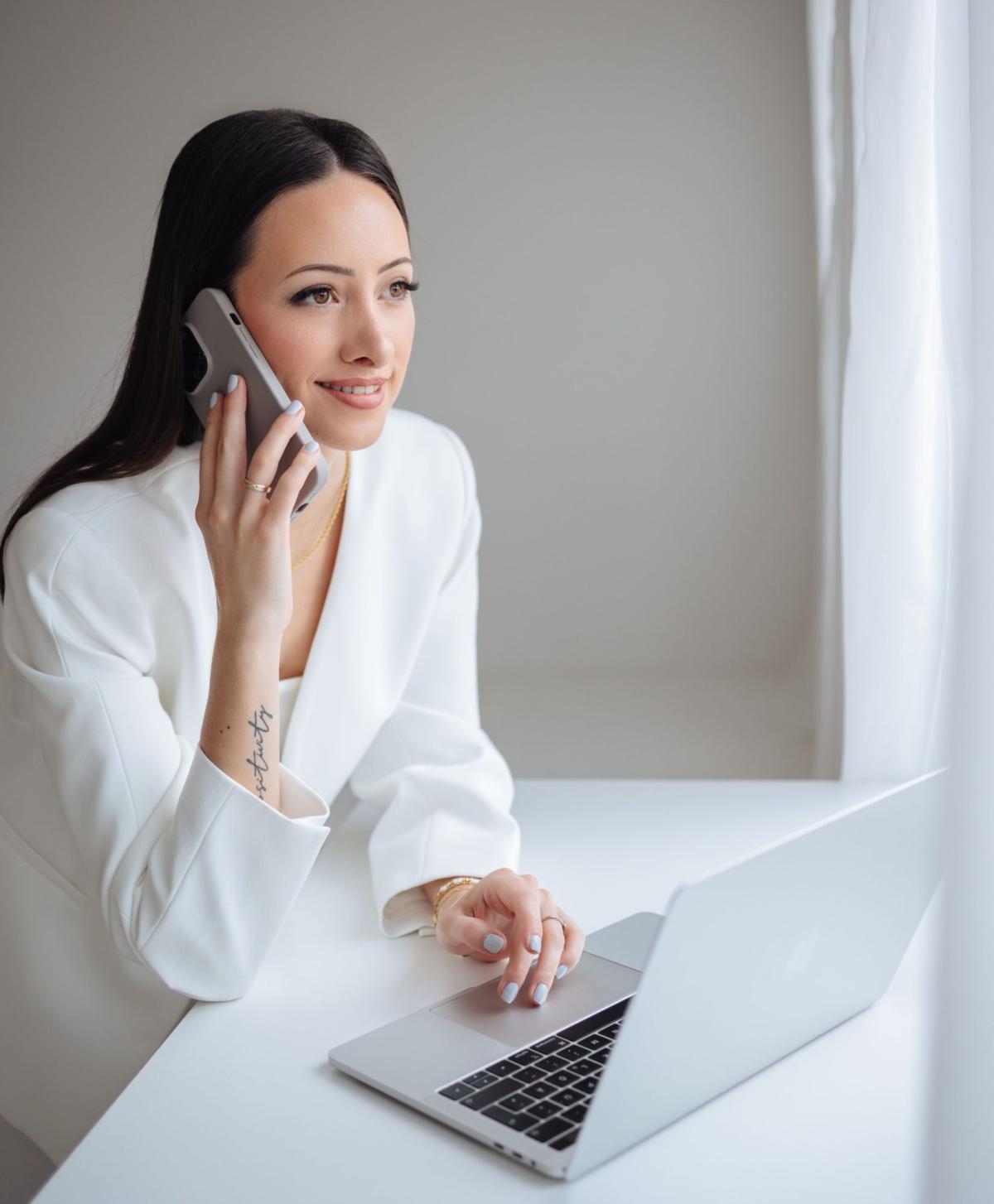 Frau im weißen Blazer spricht am Telefon und arbeitet am Laptop zum Thema Vitamin D Mangel.