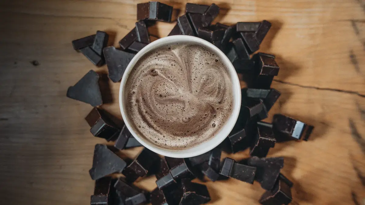 Eine Tasse zeremonieller Kakao umgeben von Schokoladenstückchen auf einem Holzuntergrund.