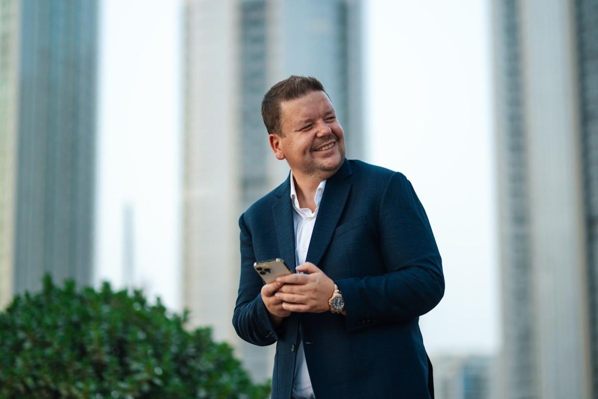 Ein Mann im dunklen Anzug hält ein Smartphone vor modernen Hochhäusern – seriöser Makler Dubai im Gespräch.
