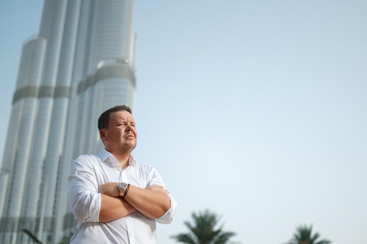 Seriöser Makler Dubai steht mit verschränkten Armen vor einem modernen Hochhaus in Dubai.