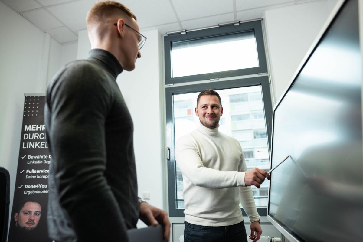 Zwei Männer diskutieren in einem Büro, während einer mit einem Pointer auf den Bildschirm zeigt.