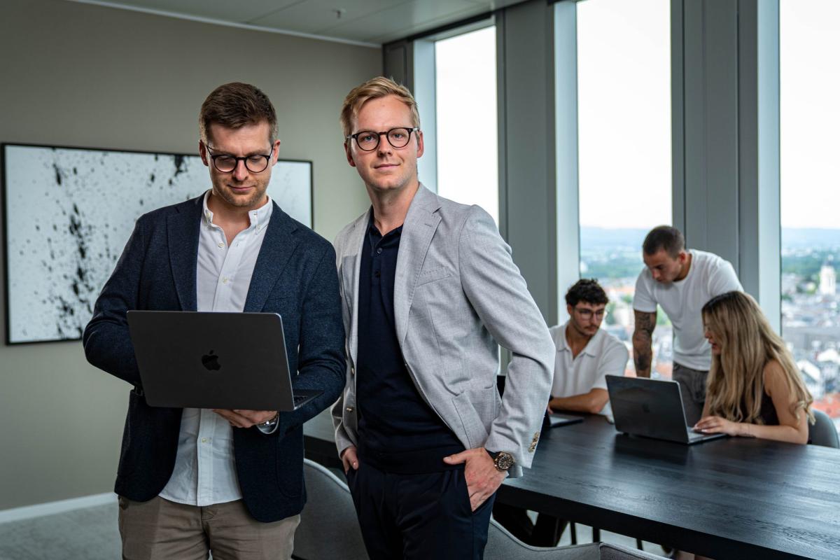 Zwei Männer im Büro präsentieren einen Laptop, während im Hintergrund ein Marketing-Team arbeitet.