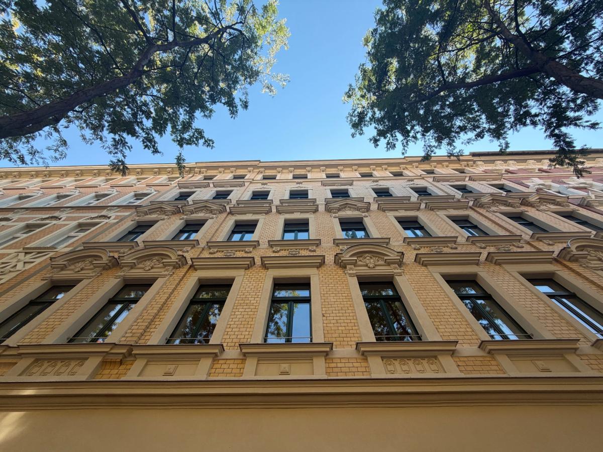 Ornamentierte Fassade einer Denkmalimmobilie, von unten fotografiert und von Baumkronen eingerahmt.