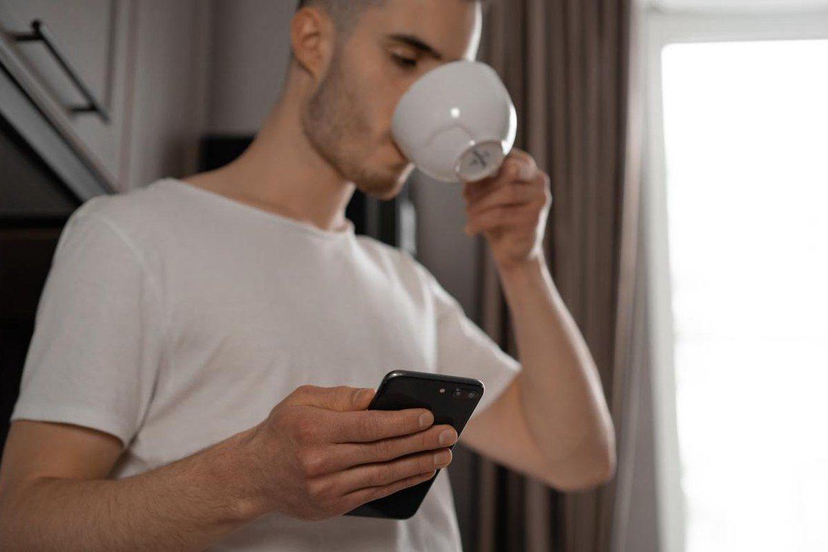 Mann mit Kaffeetasse blickt am Morgen auf sein Smartphone
