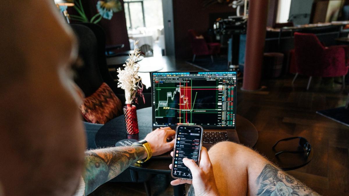 Person analysiert Trading-Daten auf Laptop und Smartphone im Wohnraum.