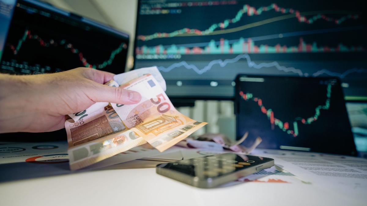 Hand hält Geldscheine über einem Handy und Finanzcharts, um Vermögen über Generationen zu schützen.