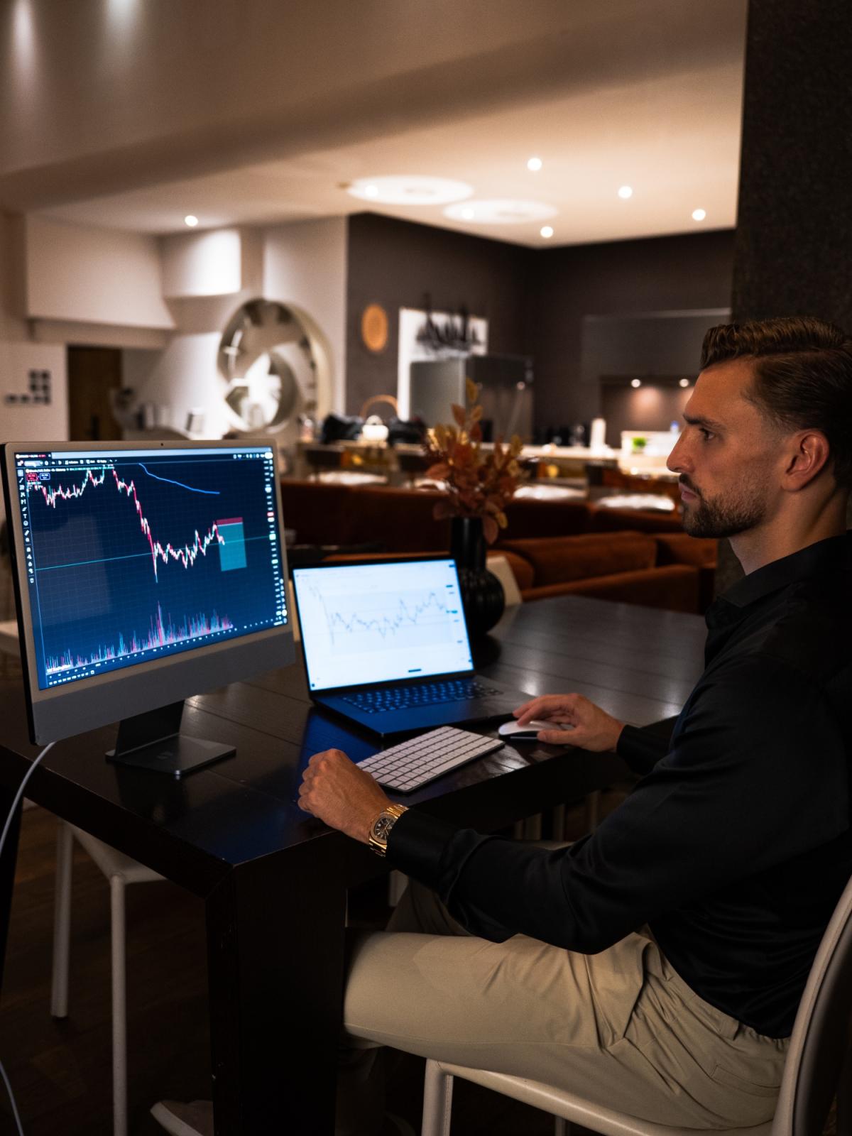 Mann analysiert Finanzcharts am Laptop und Monitor in einem modernen Raum.