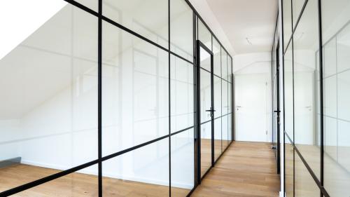 Bodentiefe Glastrennwand mit schwarzen Profilen und Drehtür trennt ein Büro vom Flur ab.