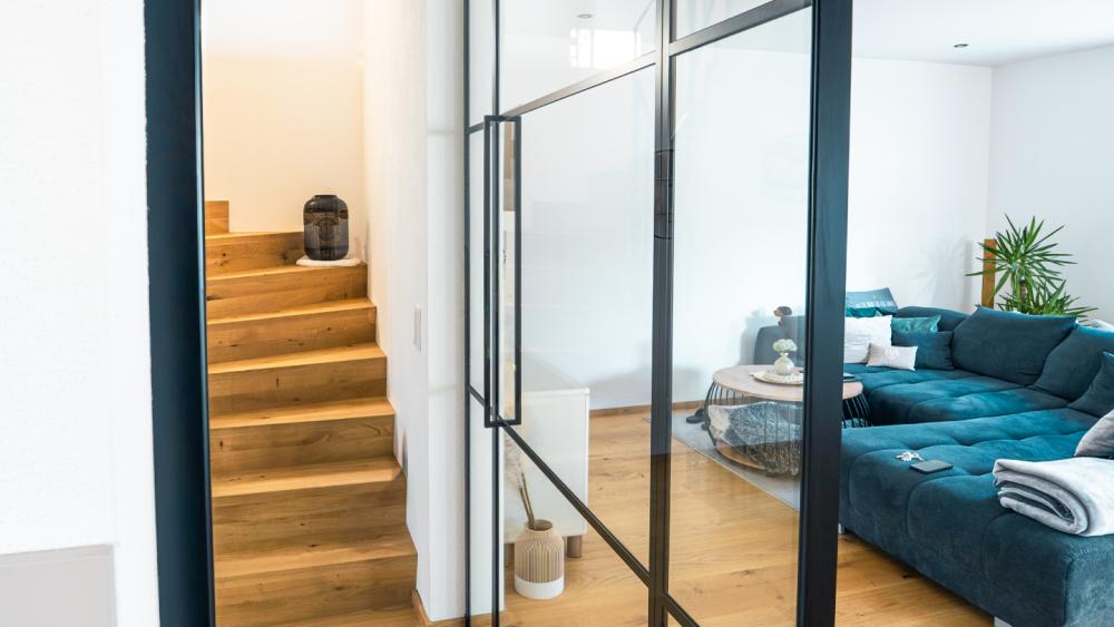 Glastrennwand Wohnzimmer mit schwarzen Metallprofilen trennt den Sitzbereich von einer beleuchteten Treppe.