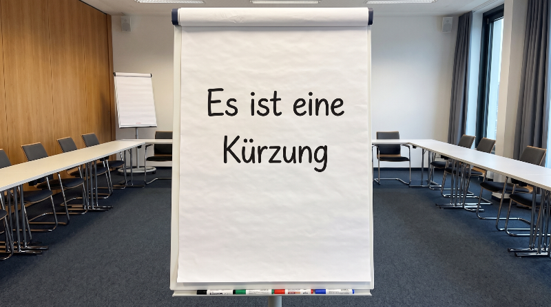 Eine Flipchart zeigt die Worte 'Es ist eine Kürzung' in einem Besprechungsraum.
