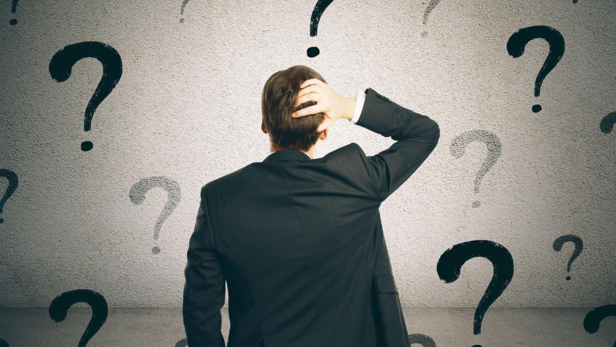 Ein Mann im Anzug steht vor einem Hintergrund mit Fragezeichen und denkt über Selbstzweifel als Unternehmer nach.