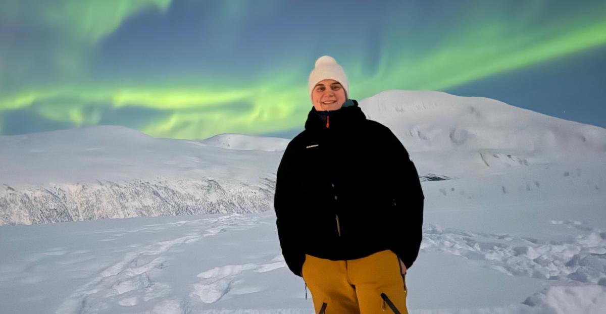Junge Person steht im Schnee unter Nordlichtern und blickt in die Kamera.