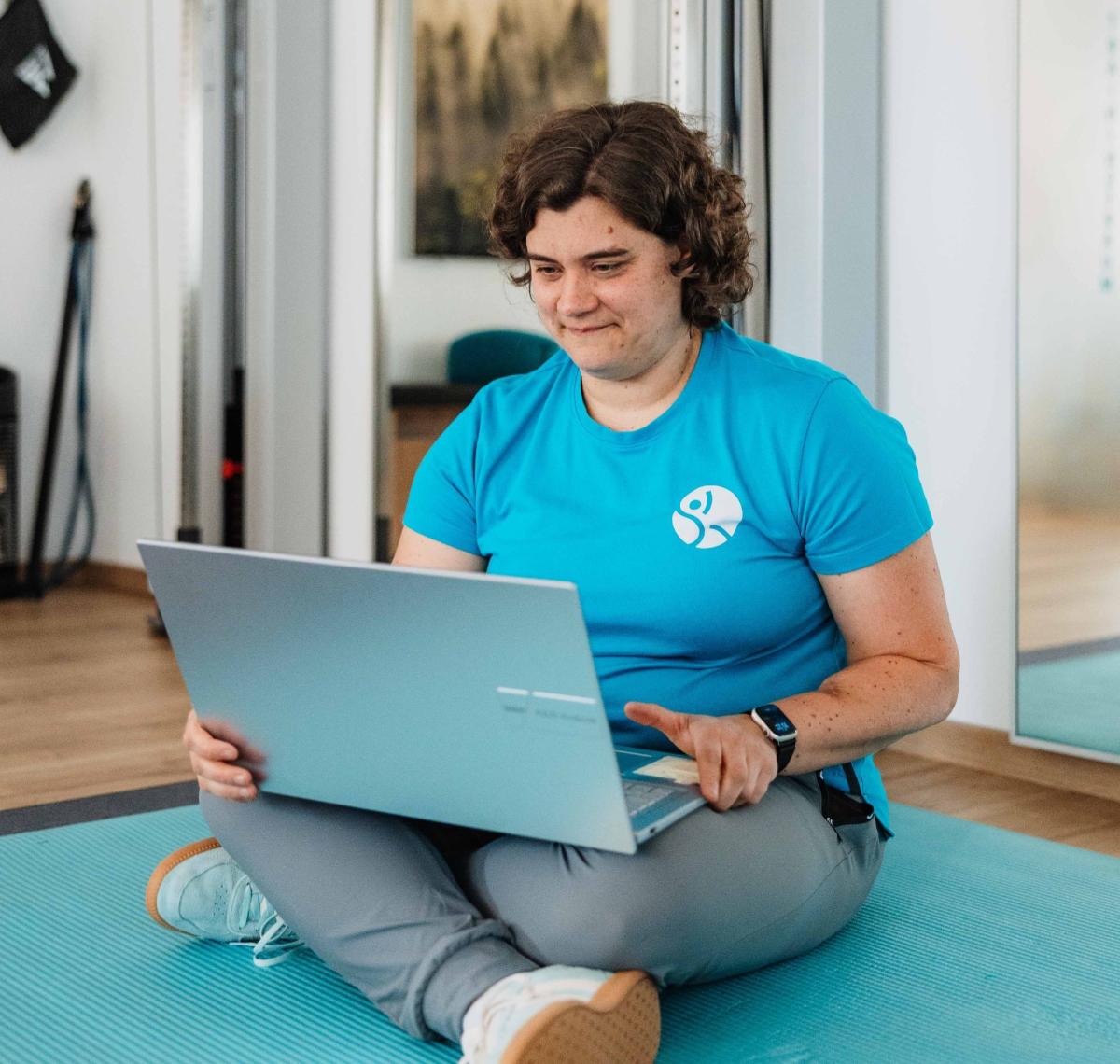 Frau sitzt auf einer Matte und arbeitet mit einem Laptop an ihrem 12-Wochen-Plan gegen Schmerzen.