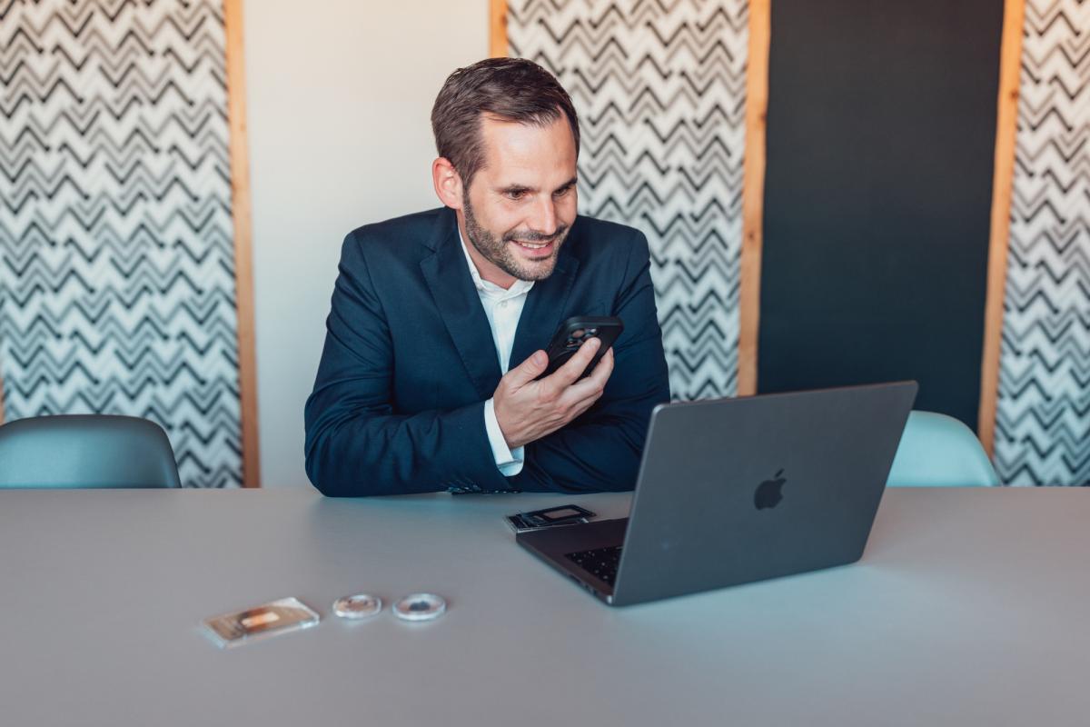 Ein Mann im Anzug sitzt am Laptop und hält ein kleines Gerät in der Hand; der Gold-Silber SWITCH steht im Mittelpunkt.
